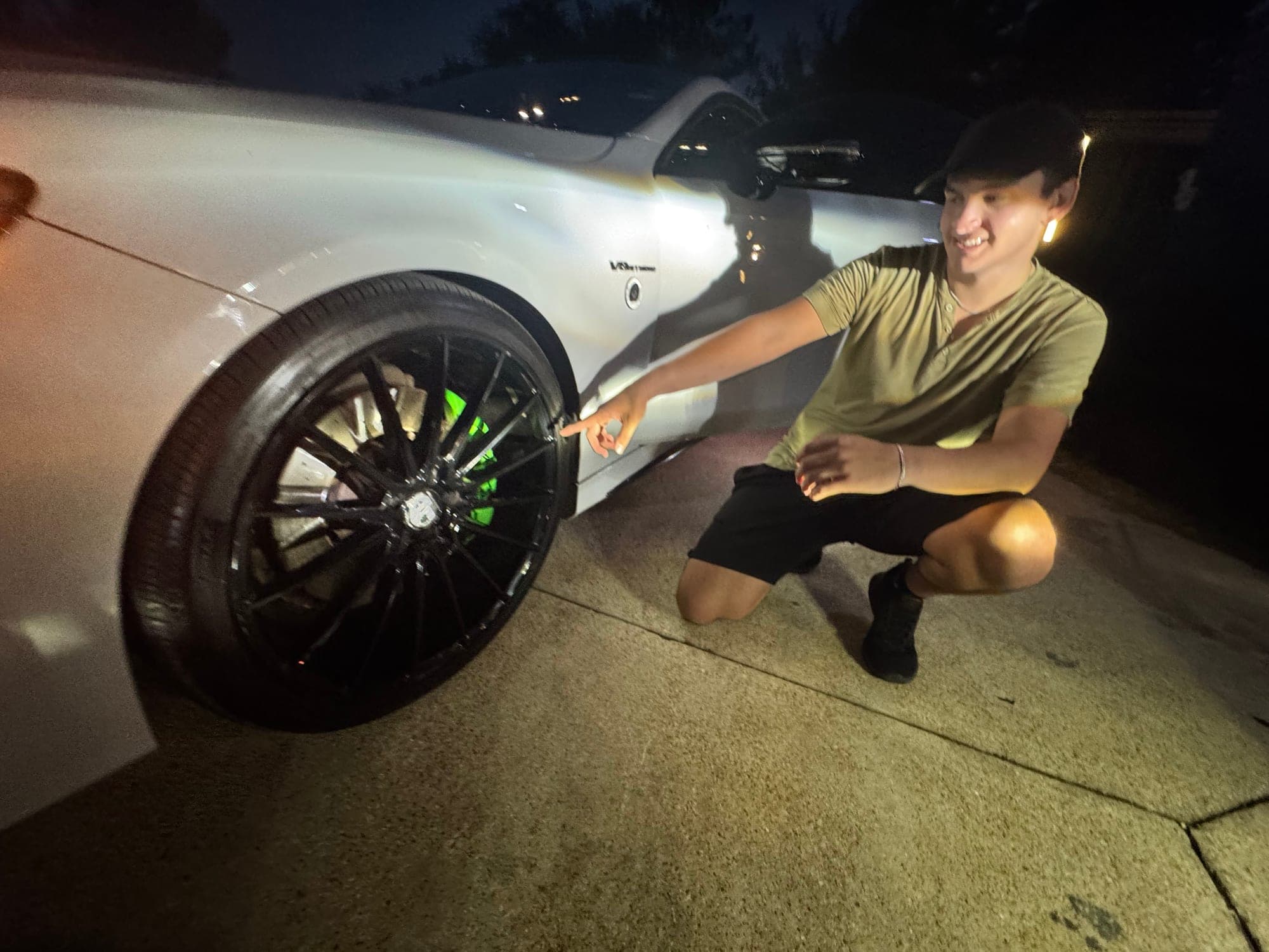 The owner of Polish Too Perfect Detailing kneeling beside a freshly detailed car at night, pointing out the finish on a wheel